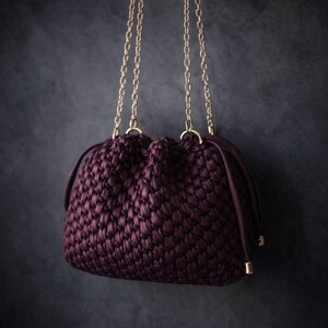 May include: A plum-colored, woven handbag with a drawstring closure and gold-tone chain straps. The bag has a textured appearance and is suspended against a dark background.