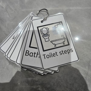 May include: A set of white laminated cards held together by a metal ring. Each card features a black line drawing of bathroom fixtures, including a toilet, shower, and bathtub, with the printed text "Bath" and "Toilet steps" on the front card.