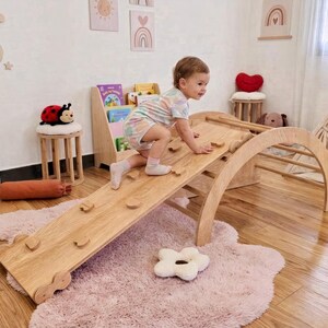 Può includere: Un set di arco e rampa per l'arrampicata in legno per bambini piccoli. Il set in legno naturale comprende un arco curvo e una rampa con prese per l'arrampicata. Un bambino sta arrampicando sulla rampa, che è posta su un tappeto rosa.