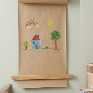 Pu&ograve; includere: Un dispenser da parete per carta kraft con un bastone in legno chiaro sostenuto da cinghie in pelle marrone. Un lungo foglio di carta marrone pende verso il basso, mostrando un colorato disegno infantile con un arcobaleno, un sole giallo, una casa blu e un albero verde.
