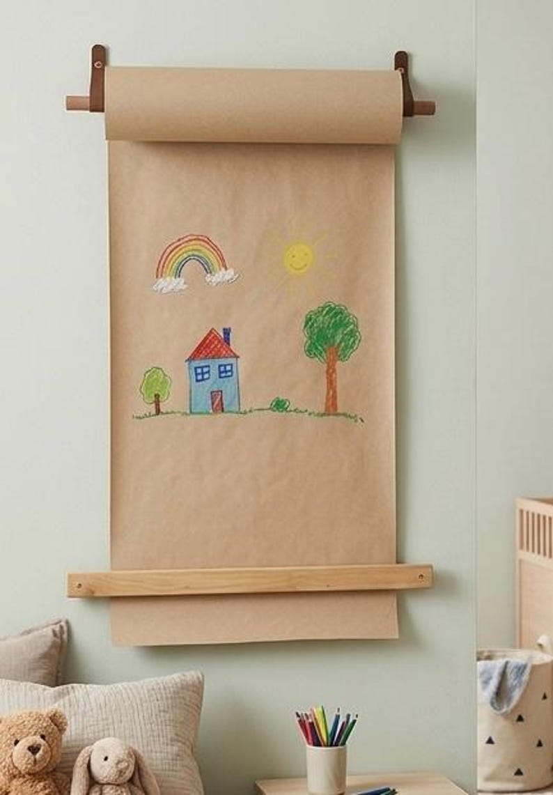 Pu&ograve; includere: Un dispenser da parete per carta kraft con un bastone in legno chiaro sostenuto da cinghie in pelle marrone. Un lungo foglio di carta marrone pende verso il basso, mostrando un colorato disegno infantile con un arcobaleno, un sole giallo, una casa blu e un albero verde.