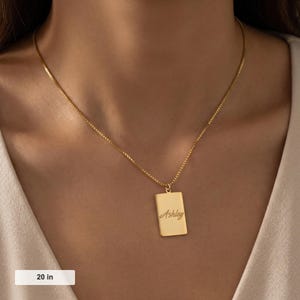 May include: A gold-colored rectangular pendant necklace with the name "Ashley" in cursive script. The chain is 20 inches long. The necklace is worn around the neck.