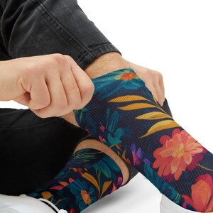Peut inclure: Une paire de chaussettes montantes avec un fond bleu marine foncé et un motif floral vibrant. Le design présente de grandes fleurs roses et orange, des feuilles bleu sarcelle et un feuillage jaune doré. Le tissu a une texture tricotée visible.