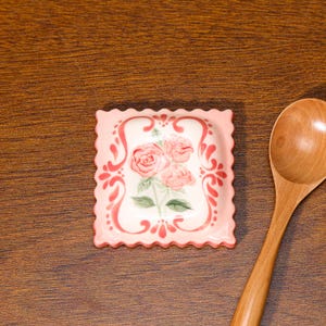 May include: A square, pink ceramic dish with a scalloped edge. The dish features a floral design of pink roses and green leaves. A wooden spoon rests on a wooden surface next to the dish.