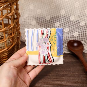 Può includere: Un piattino quadrato in ceramica con bordi smerlati, caratterizzato da un disegno dipinto a mano con strisce verticali blu, una fascia orizzontale gialla e un grappolo centrale di fiori viola su sfondo bianco con accenti rossi e neri.