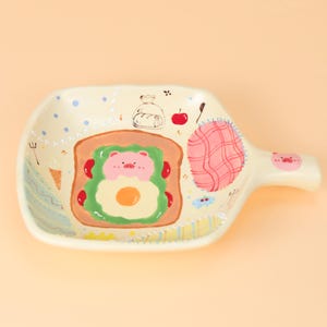 Puede incluir: Una fuente de cerámica de color crema con asa. La fuente presenta un diseño pintado a mano de un sándwich con cara de cerdo, una manzana roja y un parche a cuadros rosa. El asa tiene una cara de cerdo rosa.