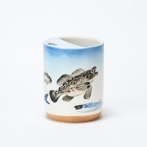 Puede incluir: Un vaso cilíndrico de cerámica con un diseño pintado a mano de varios peces en tonos marrón, gris y azul sobre un fondo blanco. El borde superior tiene un degradado azul claro y la base es de arcilla natural sin esmaltar de color tostado.