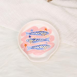 May include: A white ceramic spoon rest featuring a hand-painted design of three blue and yellow spotted fish swimming horizontally. The background includes a soft pink wash and small orange and blue star shapes scattered around the fish.