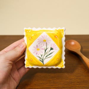 May include: A square, ceramic spoon rest with a yellow border and a white center featuring a floral design. The flowers are pink and white daisies with green stems. The spoon rest has a scalloped edge.