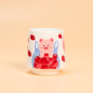 May include: White ceramic mug with a whimsical design featuring a pink pig holding a bag of red cherries. The mug has a flared base and is decorated with additional cherries. The design is playful and colorful.