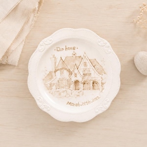 Puede incluir: Un plato redondo de cerámica blanca con borde festoneado que presenta una ilustración en tinta marrón de una casa en el centro. El texto "Our home" está impreso sobre la casa y "March 22th, 2026" debajo en letras de color marrón.