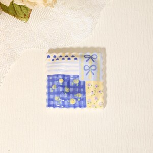 Può includere: Un piattino quadrato in ceramica con bordo smerlato, caratterizzato da un design patchwork con strisce blu e bianche, fiocchi blu su sfondo giallo pallido, un motivo a quadri blu con puntini gialli e una sezione gialla con piccoli fiori viola.