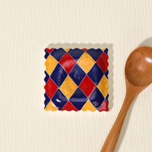 Puede incluir: Un plato cuadrado de cerámica con un patrón de arlequín en rombos rojos, amarillos y azul marino. El plato tiene un borde festoneado. Una cuchara de madera está en el fondo.