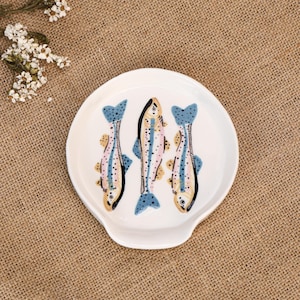 Può includere: Un poggia cucchiaio in ceramica bianca con un'illustrazione dipinta a mano di tre trote disposte verticalmente. I pesci sono raffigurati in tonalità di blu, rosa e giallo con macchie nere sulla superficie liscia e lucida del piattino.