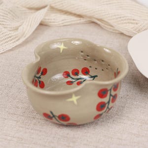 May include: A beige ceramic berry bowl with a scalloped rim and a series of small drainage holes near the top. The bowl features a hand-painted pattern of red cherry clusters on green stems and small yellow four-pointed stars on the exterior.