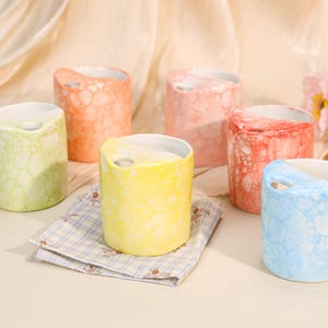 Może przedstawiać: Kolekcja sześciu ceramicznych kubków w różnych pastelowych kolorach, w tym zielonym, pomarańczowym, różowym, czerwonym, żółtym i niebieskim. Każdy kubek ma unikalną bąbelkową teksturę na zewnątrz, białe wnętrze i mały otwór przy krawędzi.