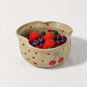 May include: A beige ceramic berry bowl with a scalloped rim and drainage holes on the side. The exterior features hand-painted red cherries and small red polka dots. The bowl is filled with fresh red strawberries and dark blue blueberries.
