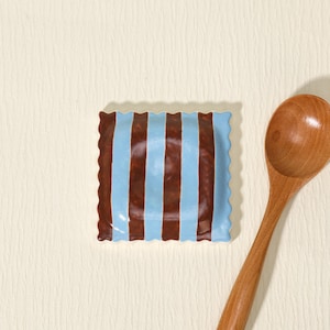 May include: A square, ceramic dish with a wavy edge, featuring alternating vertical stripes of light blue and brown. A wooden spoon rests beside the dish on a cream-colored surface. The dish is likely for serving food.