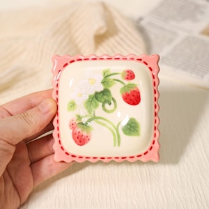 Pode incluir: Um prato quadrado de cerâmica com uma borda rosa ondulada e um desenho de morango pintado à mão. O prato apresenta morangos vermelhos, flores brancas e folhas verdes sobre um fundo creme. O prato é segurado por uma mão.