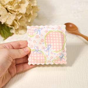 May include: A square ceramic dish with scalloped edges featuring a pastel pink gingham pattern, small floral motifs, and light blue ribbon illustrations. The surface is decorated with tiny white raised dots throughout the design.