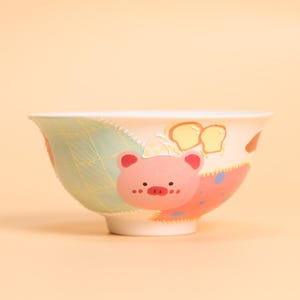 Puede incluir: Un cuenco de cerámica blanco con un diseño caprichoso. El cuenco presenta una ilustración de cerdo rosa con un gorro de chef, pan y un patrón de patchwork en tonos verdes y rosas. El cuenco es adecuado para servir alimentos.