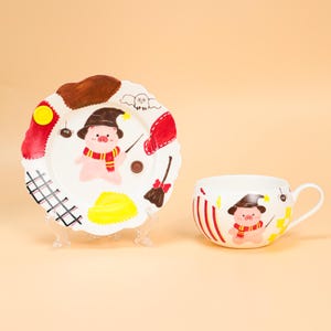 May include: A white ceramic plate and mug set featuring a cartoon pig in a wizard hat and scarf. The plate has a scalloped edge with red, yellow, and brown accents. The mug has red stripes and a handle.