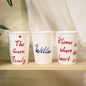 May include: Three white ceramic mugs with handwritten text in red and blue. The mugs have the phrases "The Green Family", "Willow", and "Home is where the heart is". A small red heart is above "The Green Family".