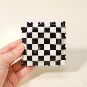 May include: A square, ceramic dish with a black and white chequered pattern. The dish has a wavy, scalloped edge. The black squares are evenly spaced against the white squares. The dish is held by a hand.