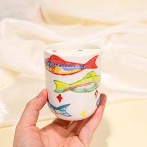 Puede incluir: Un vaso de cerámica blanca con un patrón pintado a mano de peces coloridos en tonos rojo, amarillo, verde y azul. El diseño incluye pequeñas formas de estrellas y diamantes dispersas entre las ilustraciones de peces en la superficie.