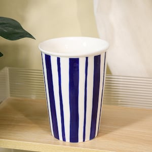 Könnte beinhalten: Ein weißer Keramikbecher mit vertikalen marineblauen Streifen. Das Innere des Bechers ist weiß. Der Becher ist zylindrisch mit einem leicht ausgestellten oberen Rand. Der Becher steht auf einer hellen Holzoberfläche.