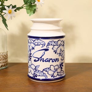 Könnte beinhalten: Eine weiße Keramikvase mit blauem Blumendesign und dem Namen "Sharon" in Schreibschrift. Die Vase hat die Form eines Milchkrugs mit breitem Boden und schmalerem Oberteil. Das Blumenmuster umfasst Blumen und Blätter. Die Vase steht auf einer Holzoberfläche.