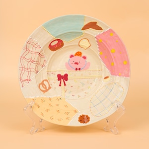 Puede incluir: Un plato decorativo de cerámica con un diseño de patchwork. El plato presenta una imagen central de un cerdo con pajarita y sombrero. Varias ilustraciones de alimentos, incluyendo un donut y un pretzel, están pintadas alrededor del cerdo. El plato está sostenido por un soporte de plástico transparente.