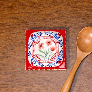 May include: Square ceramic coaster with a red scalloped edge. The center features a floral design with red flowers, green stems, and blue and white decorative elements. A wooden spoon rests nearby.