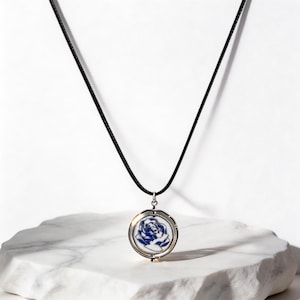 Puede incluir: Collar con colgante redondo plateado con un diseño de rosa azul sobre fondo blanco. El colgante cuelga de un cordón negro. El collar se exhibe sobre una superficie de mármol blanco.