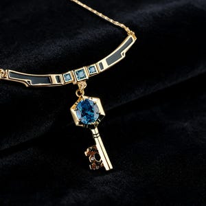 Op de afbeelding: Een gouden ketting met een sleutelvormige hanger met een centrale ovale blauwe edelsteen in een zeshoekig frame. De ketting bevat gebogen gouden segmenten met zwarte emaille en kleine vierkante blauwe edelstenen langs de halslijn.