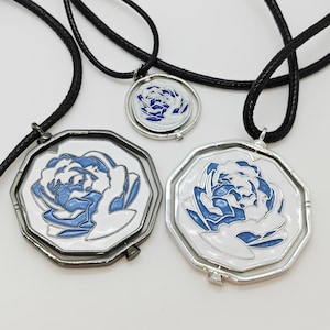 Puede incluir: Tres collares con cordones negros. Los colgantes presentan un fondo blanco con un diseño floral azul. Los colgantes tienen marcos metálicos plateados y color cañón. Los collares se exhiben sobre una superficie blanca.