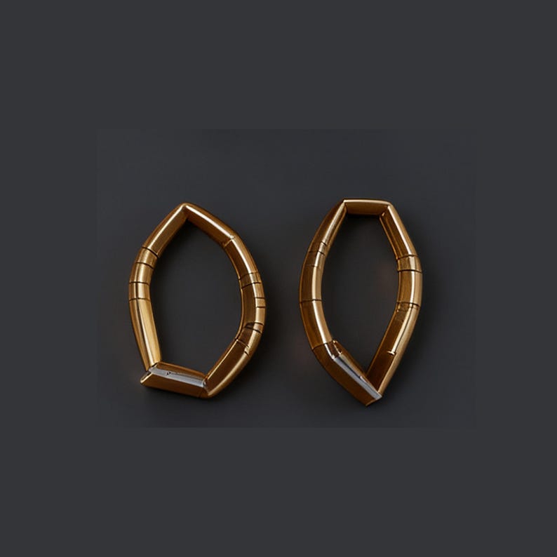 Puede incluir: Dos pendientes dorados de forma geom&eacute;trica, con forma de hoja. Cada pendiente tiene un dise&ntilde;o hueco y angular con una serie de l&iacute;neas paralelas. Se presentan sobre un fondo gris oscuro.