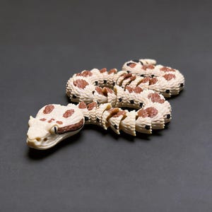Puede incluir: Un juguete de serpiente articulada con un color base blanco hueso, manchas marrones y puntos negros a lo largo de su cuerpo segmentado. La serpiente tiene una cabeza detallada con ojos visibles y una superficie texturizada con uniones.