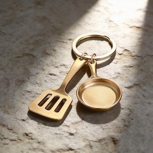 May include: A gold-toned metal keyring featuring two miniature kitchen utensil charms: a slotted spatula and a small round frying pan. The charms are attached to a circular split ring, all finished with a smooth, brushed metallic texture.