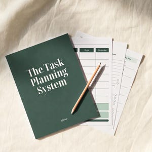 Może przedstawiać: Ciemnozielony notatnik z napisem "The Task Planning System" w białych literach. Obok notatnika znajduje się kilka kartek papieru i drewniany ołówek. Kartki mają siatkę i linie do pisania. Tło to jasnobeżowa tkanina.