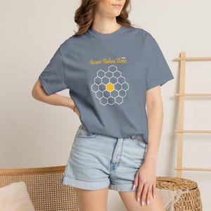 May include: A blue t-shirt with the text "Sweet Takes Time" in gold script above a honeycomb design. The honeycomb is white with a yellow center. The shirt is a solid color and has short sleeves.