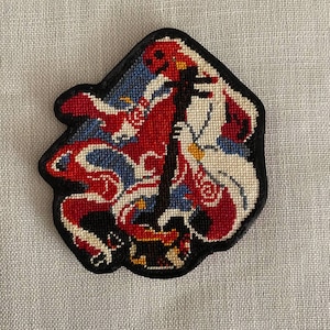 Könnte beinhalten: Ein Nadelpatch mit schwarzem Rand. Der Patch zeigt ein Design mit roten, weißen und blauen Farben. Das Design scheint eine stilisierte Figur oder ein Emblem zu sein. Der Hintergrund ist ein hellgrauer Stoff.