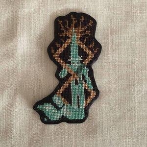 May include: A cross-stitch patch with a teal, brown, and black design. The patch has a dark brown border and features a geometric pattern with a central teal element. The background is a neutral linen fabric.