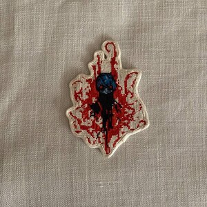 May include: A cross-stitched patch featuring a stylized figure with blue hair and a black outfit, set against a backdrop of red, blood-like patterns. The patch has a white border and is likely for clothing or accessories.