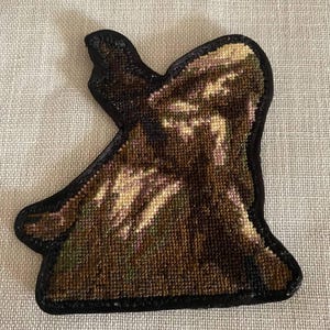 May include: A handcrafted patch with a black border and a detailed, textured design. The patch features a mix of brown, beige, and green threads, creating a camouflage-like pattern. The patch is likely for clothing or accessories.