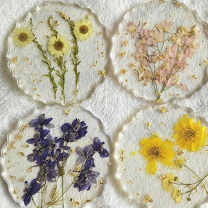 May include: Four circular resin coasters with scalloped edges, each containing pressed flowers and gold leaf flakes. One features yellow daisies, another pink larkspur, the third purple delphinium, and the fourth bright yellow wildflowers.
