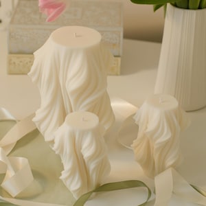 Könnte beinhalten: Elfenbeinfarbene Kerzen mit einem Design aus drapiertem Stoff. Die Kerzen sind in verschiedenen Größen erhältlich und werden mit dekorativen Bändern und einer weißen Vase arrangiert. Die Kerzen haben ein einzigartiges, künstlerisches Aussehen, das sich für die Inneneinrichtung eignet.