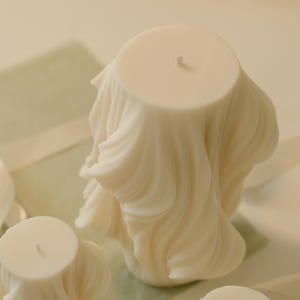 May include: Ivory-coloured pillar candles with a draped fabric design. The candles have a smooth, waxy texture and a wick at the top. The background includes a pale green surface and a white ribbon.