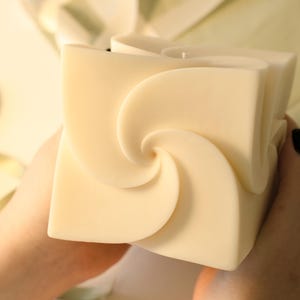 Könnte beinhalten: Eine große, quadratische Kerze in cremefarbenem Off-White. Die Kerze hat ein einzigartiges Spiralmuster auf jeder Seite, das ein visuelles Interesse erzeugt. Die unparfümierte Kerze ist ein dekoratives Element für das Zuhause.