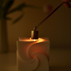 Könnte beinhalten: Eine brennende weiße Kerze mit einem einzigartigen Spiraldesign. Eine Hand hält einen Messing-Kerzenlöscher über der Flamme. Die Kerze befindet sich in einem schwach beleuchteten Raum und erzeugt eine warme Atmosphäre.